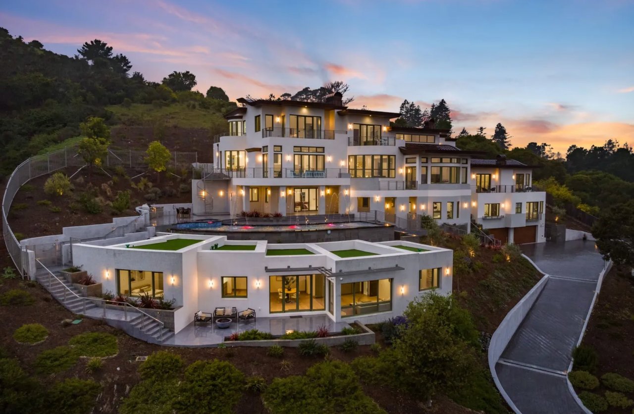 A sprawling modern mansion with a pool and expansive views sits atop a hillside.