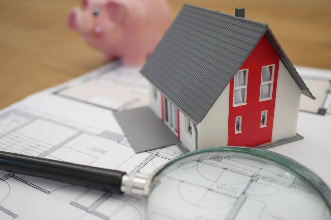A small model house with a red and white exterior and a gray roof sits on top of architectural blueprints, symbolizing real estate or home planning.
