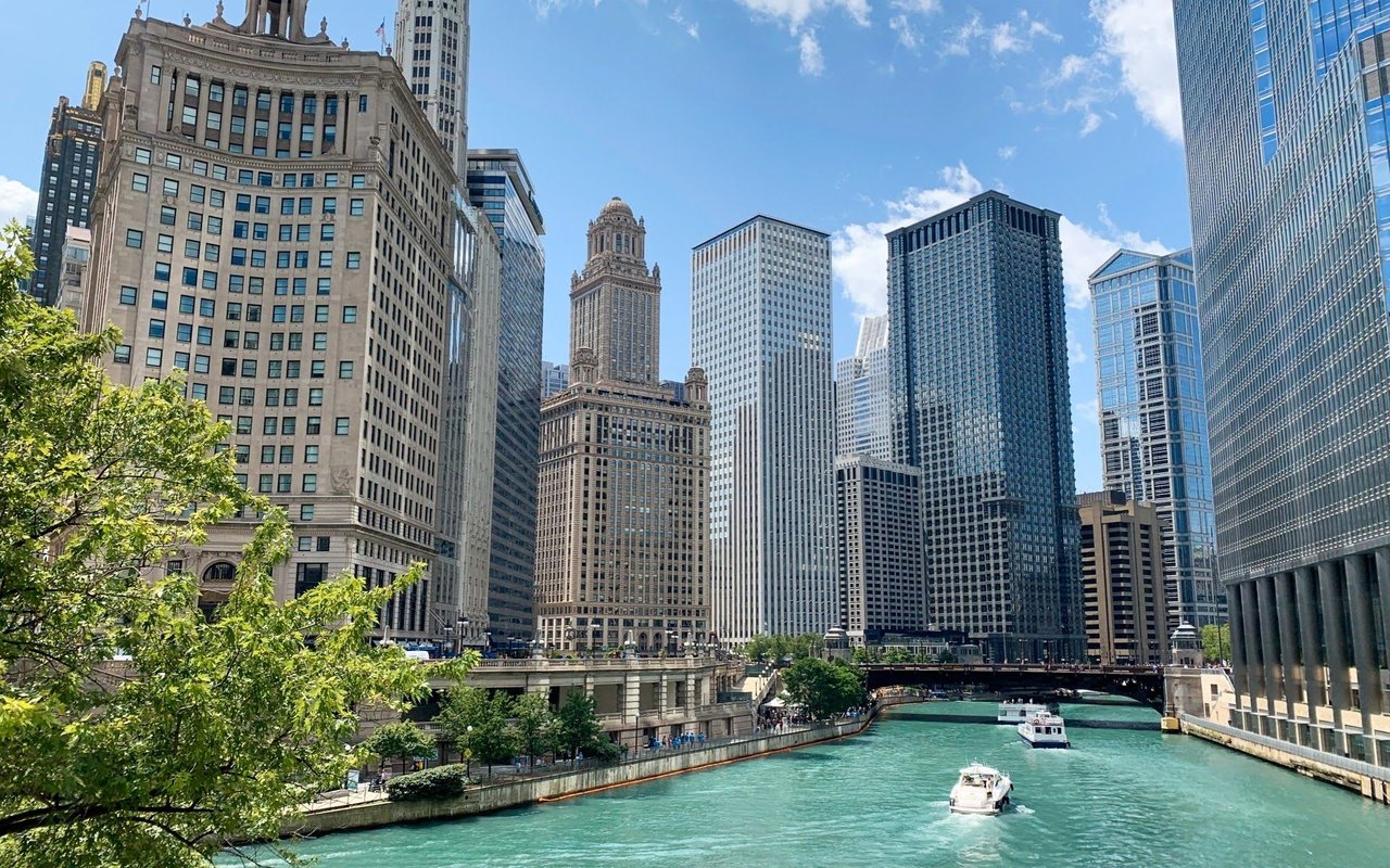 A river flows through the city, flanked by skyscrapers, with a bridge spanning overhead and boats dotting the water.