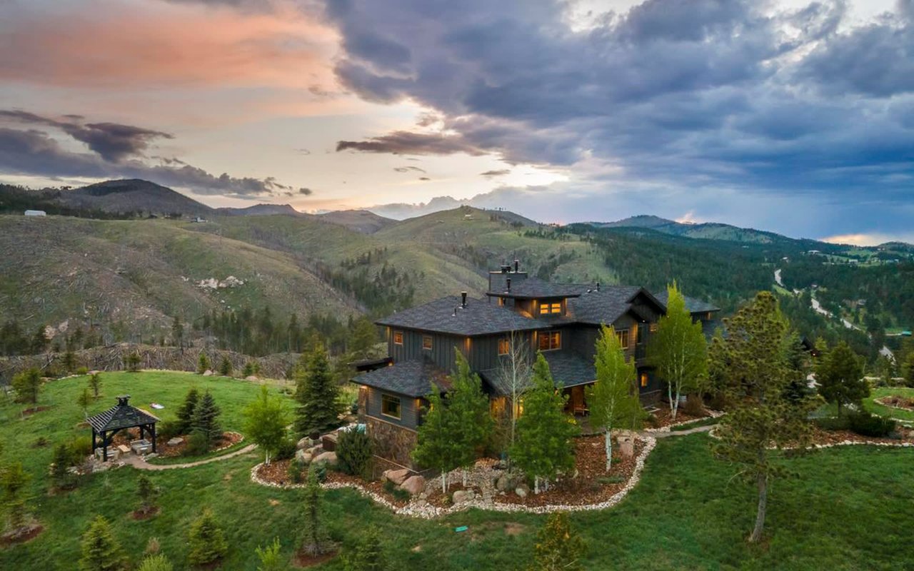 A large house perched on top of a hill. In front of the house, there is a stone patio with a wooden bench and a fire pit.