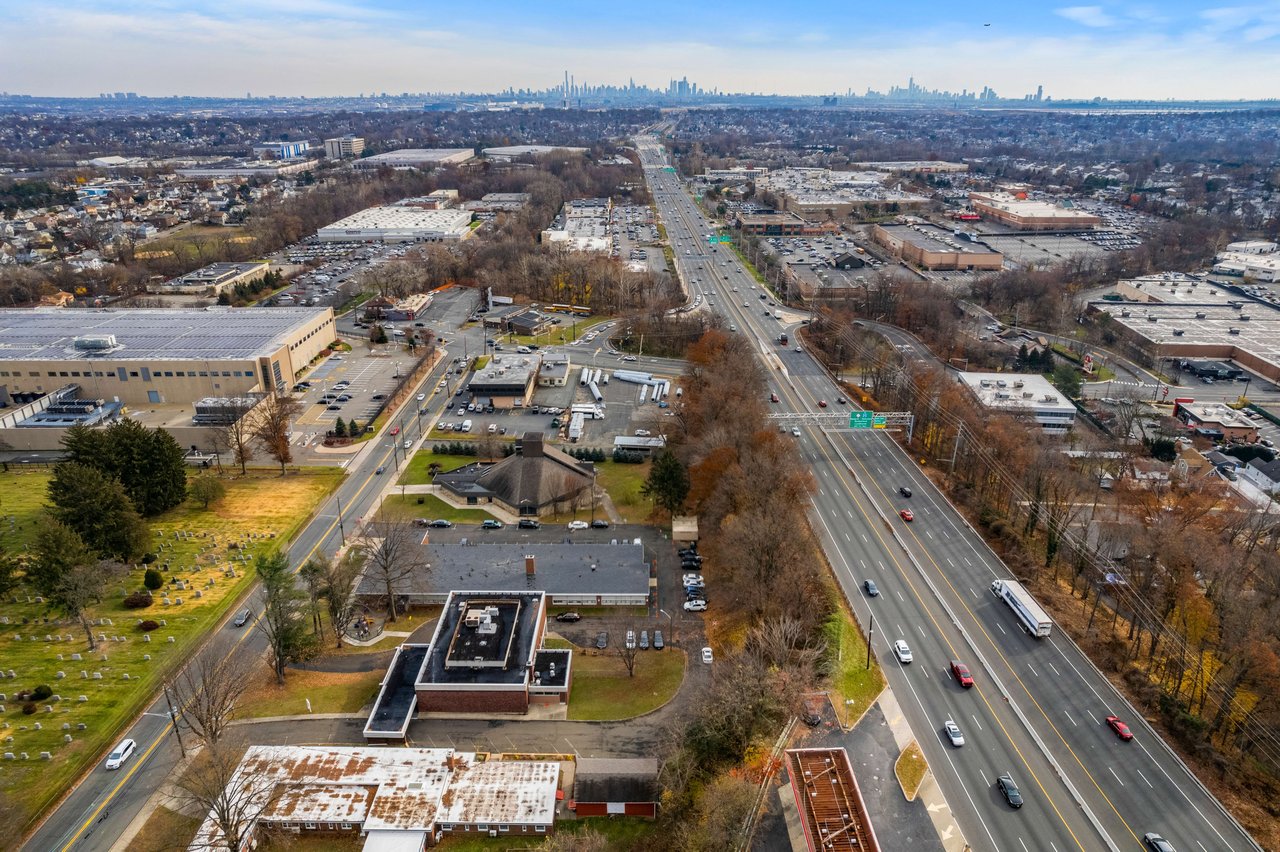 An aerial view of a highway interchange and a surrounding city, with buildings, trees, and green space.