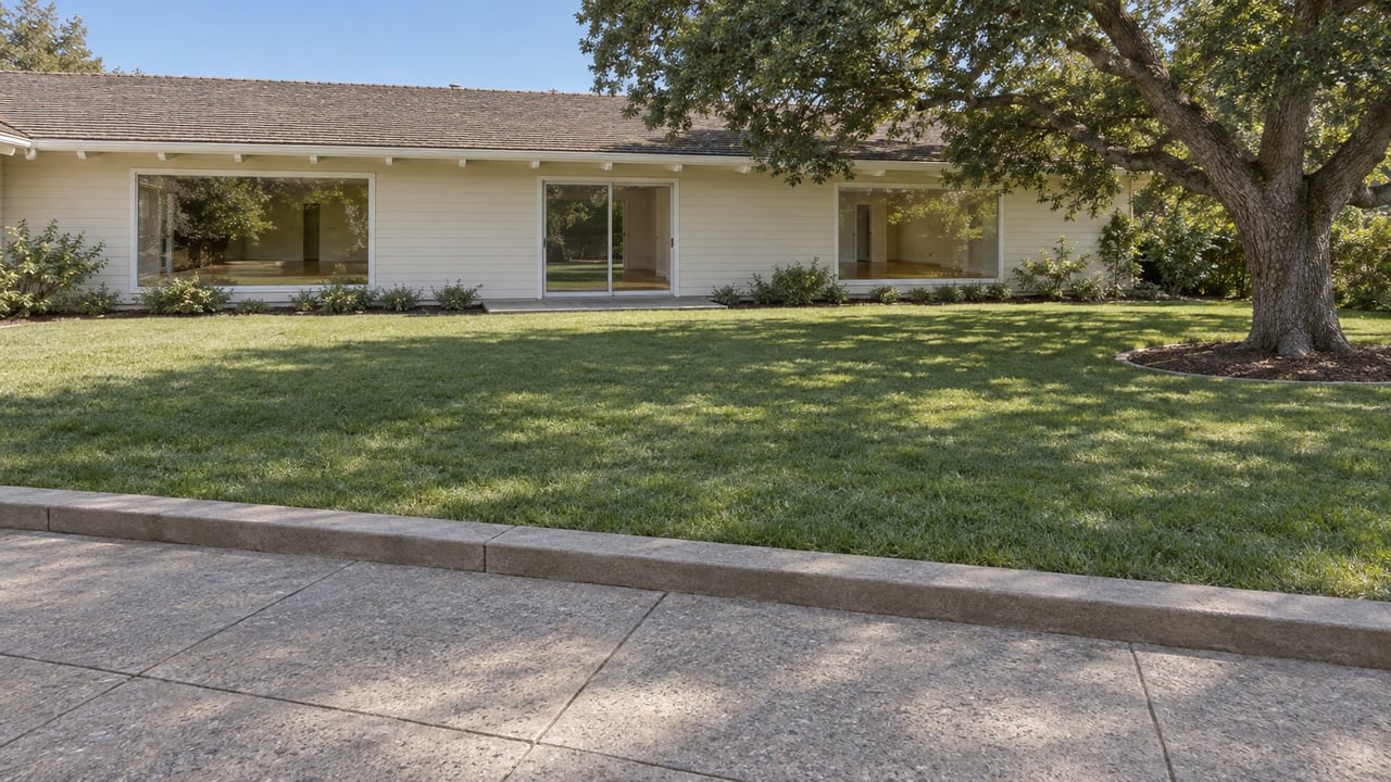 An ivory-sided ranch home across a broad lawn, viewed from a concrete terrace beside a mature spreading oak.