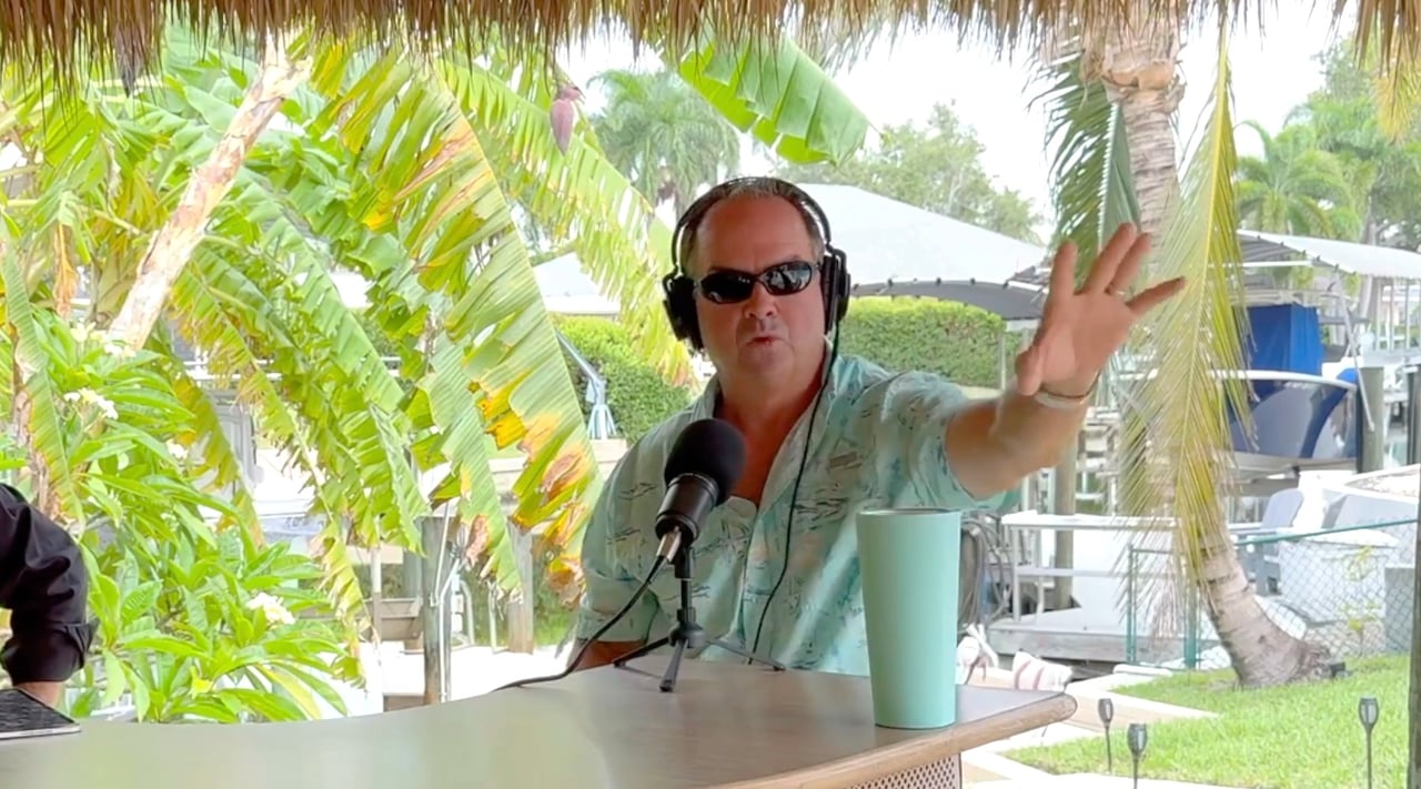 Man in sunglasses and headphones gestures beside a microphone in a tropical waterfront setting.