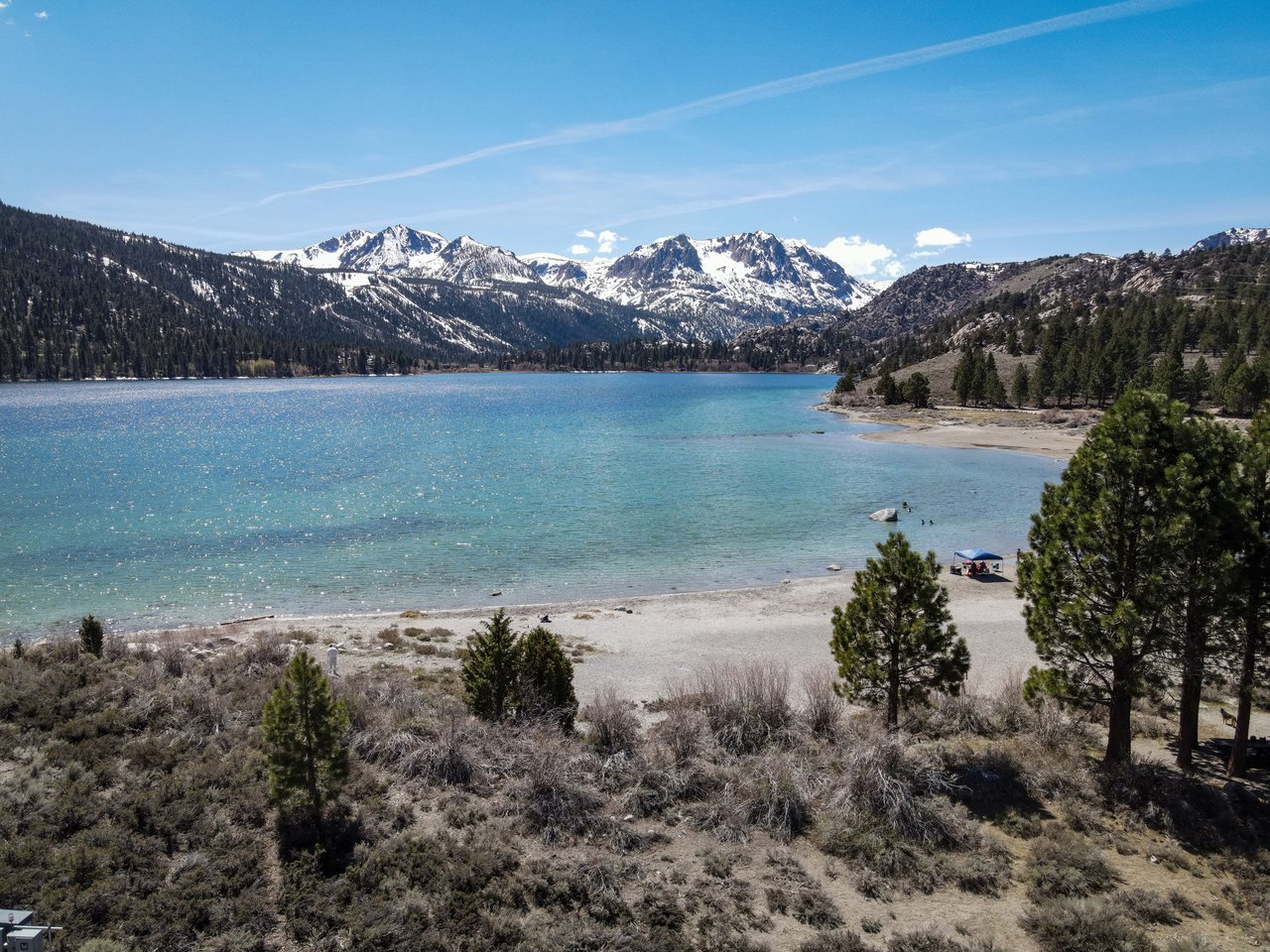 Turquoise alpine lake below snow-capped mountains, with a sandy shore and pine-covered hills.