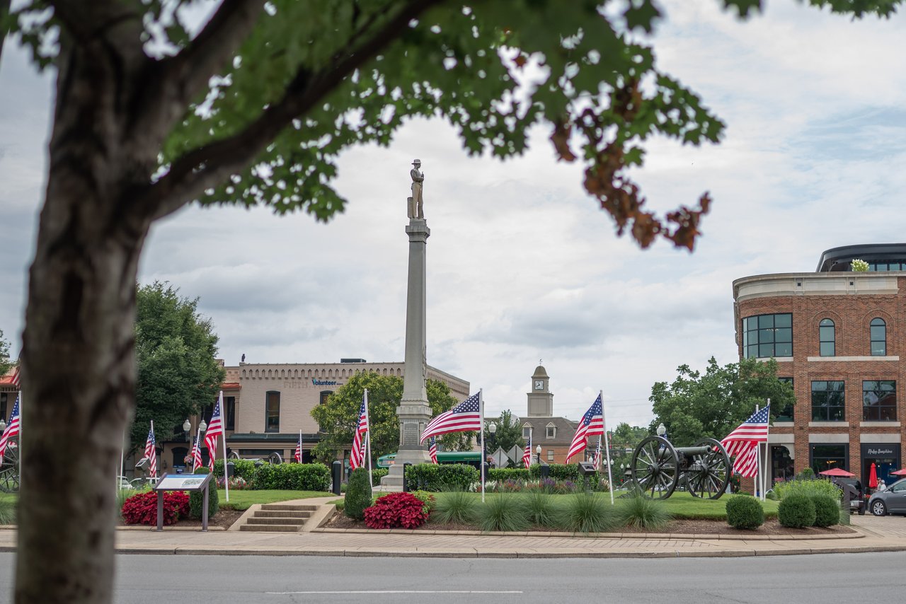Town square memorial with a statue-topped column, American flags, and historic cannons.