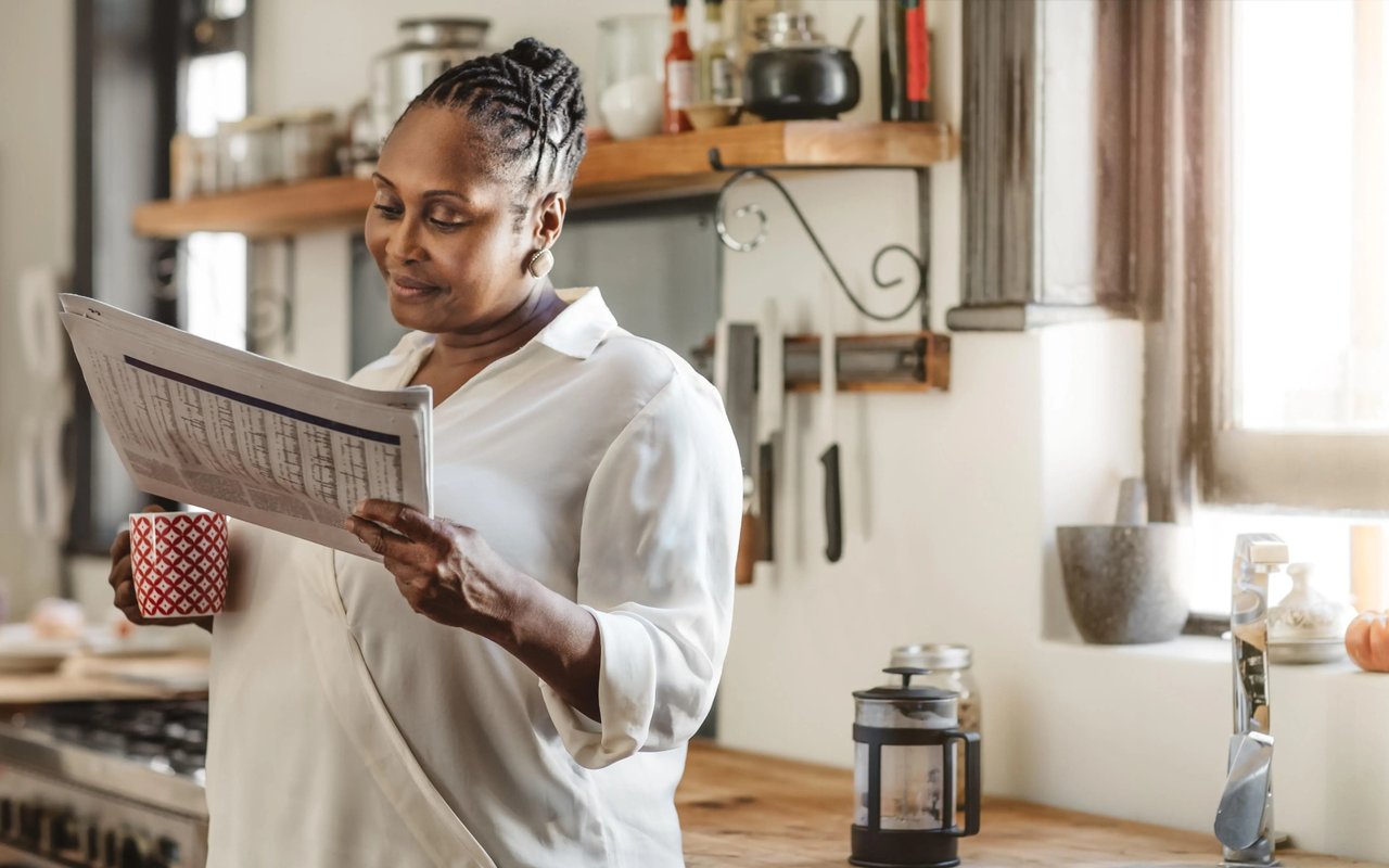 Woman reading a newspaper and holding a patterned mug in a bright kitchen.