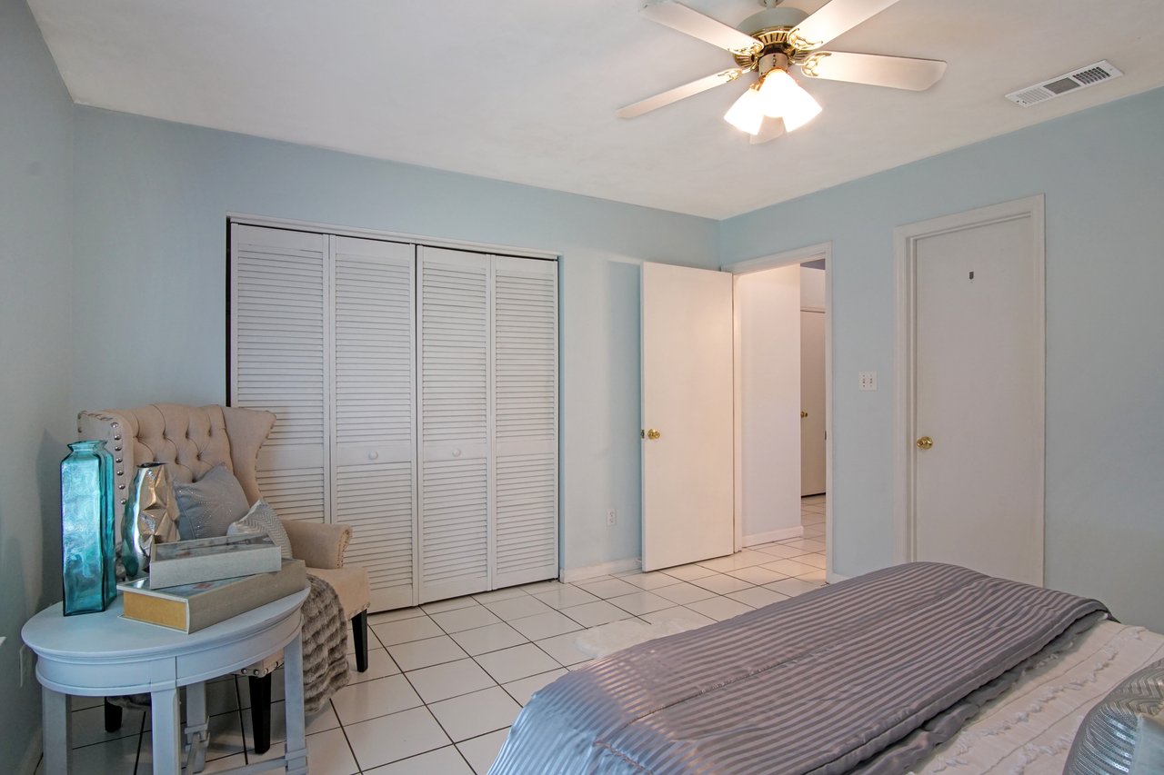 Spacious bedroom with soft blue walls, white tiled floor, and a bed with striped gray bedding. There's a cushioned chair with decorative pillows, a ceiling fan, and a closed closet with louvered doors, creating a serene and modern atmosphere.