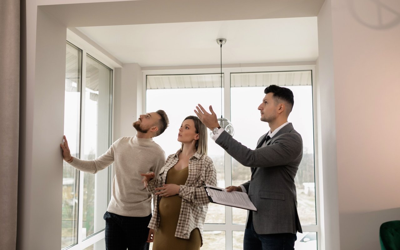 Buyers view a bright, empty room with large windows as an agent explains.