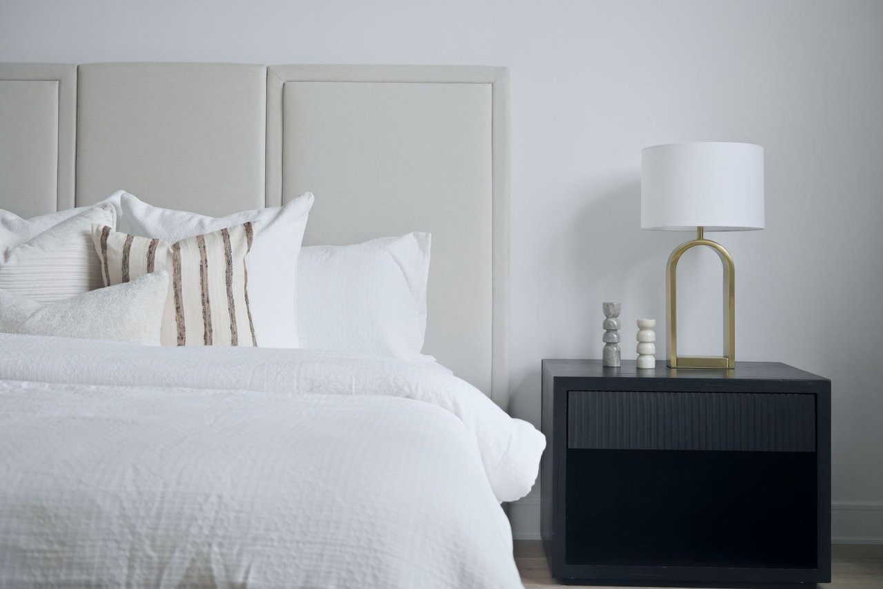 Staged bedroom with a white bed, striped accent pillow, upholstered headboard, and brass lamp.