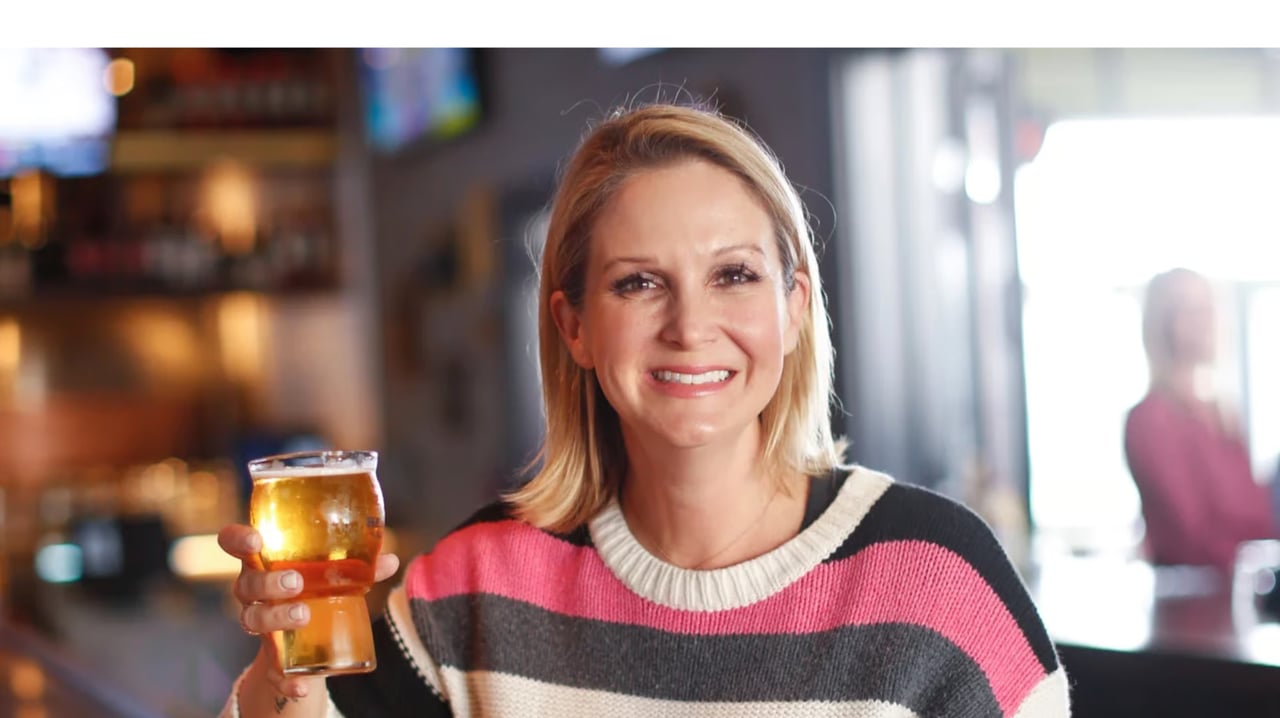 A blonde woman smiling at the camera, holding a glass of beer in a bar with a blurred background.