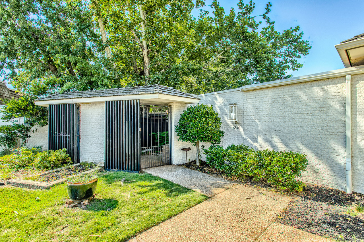 Home entrance with a black barred gate, white brick walls, and a walkway bordered by shrubs.