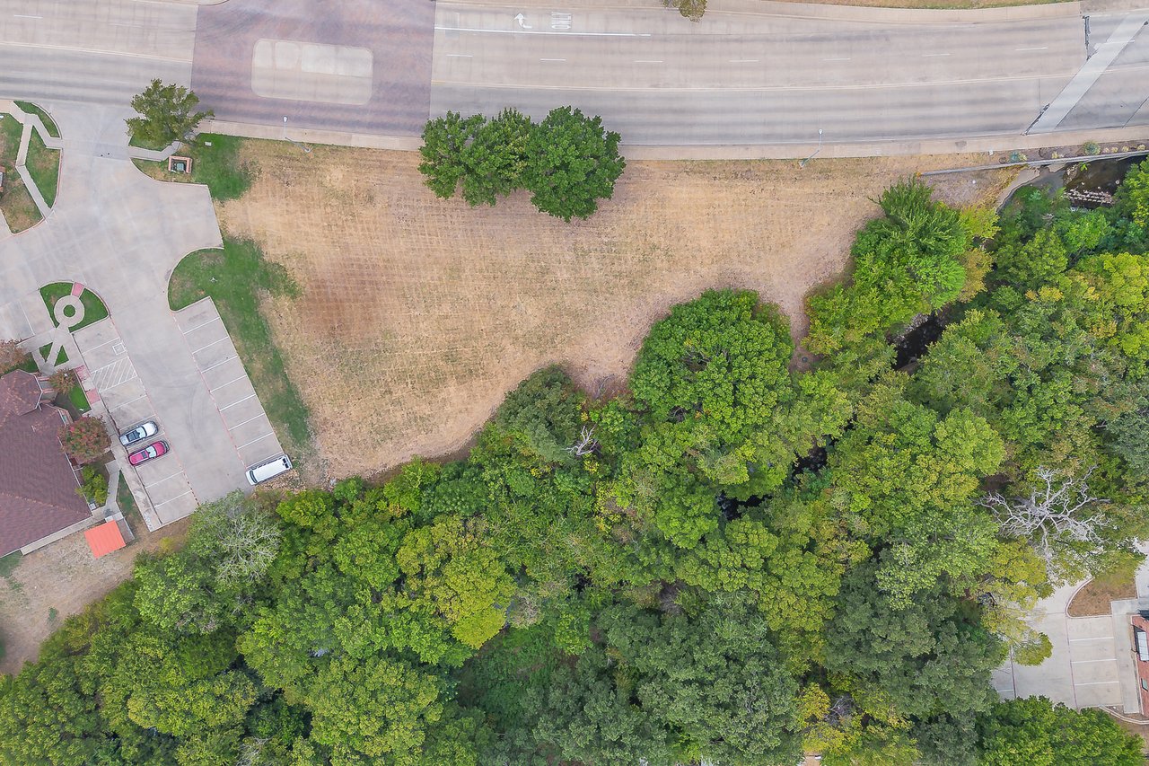 Aerial view of a vacant lot beside a road, parking area, and mature trees.