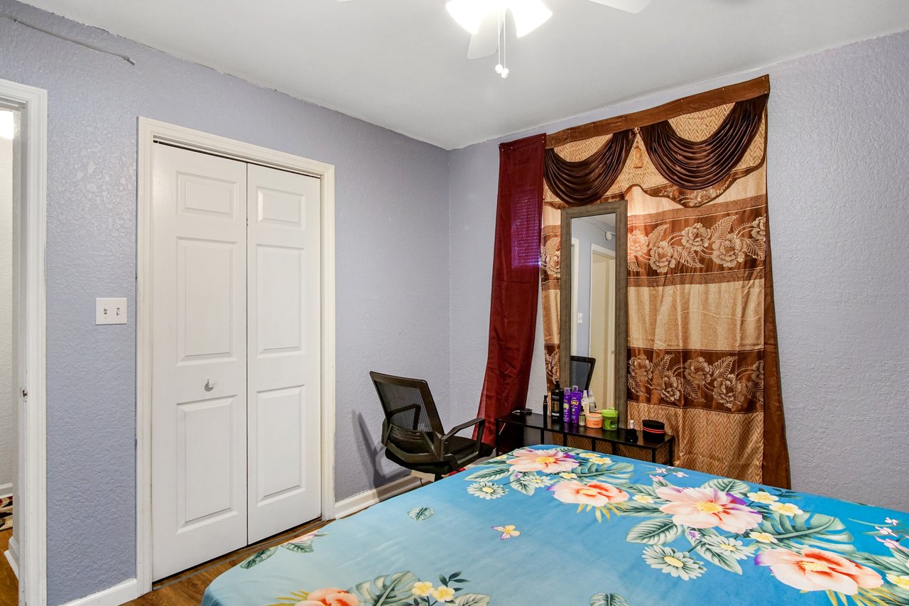 Bedroom at 2506 Nugget Lane in Tallahassee, Florida, featuring lavender-painted walls, ceiling fan with light, and wood-look flooring. The room includes a double-door closet, a small desk with chair and mirror, and a window with decorative curtains.