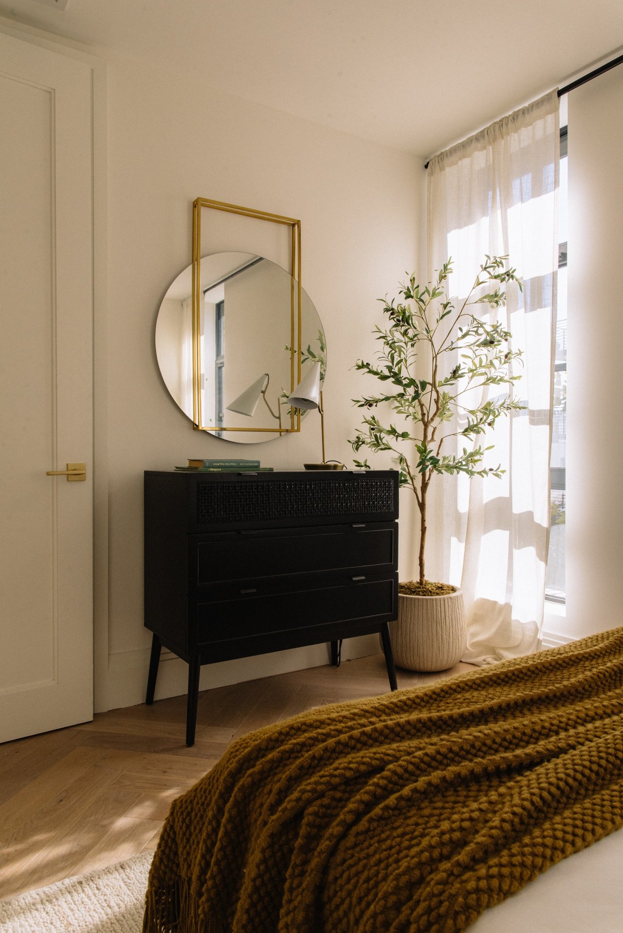 Bedroom with a chunky knit blanket, black dresser, layered mirrors, and potted tree beside a bright window.