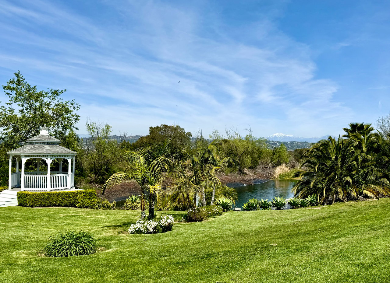 Landscaped grounds with a white gazebo, pond, palm trees, and distant snow-capped mountains.