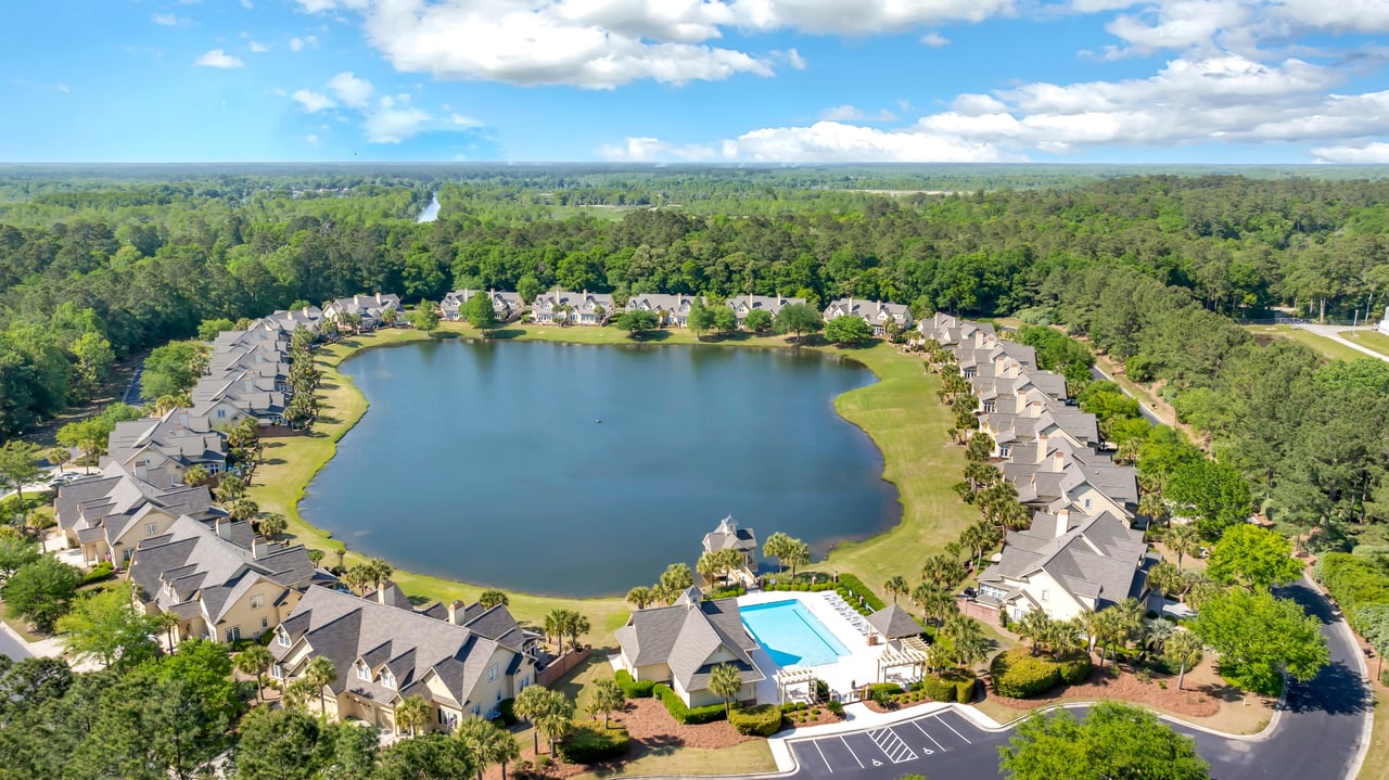 Aerial view of homes surrounding a lake, with a swimming pool and forest beyond.