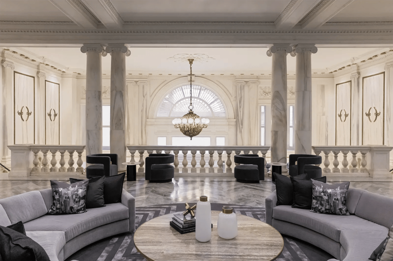 Furnished lobby lounge with marble columns, a chandelier, and curved gray sofas.