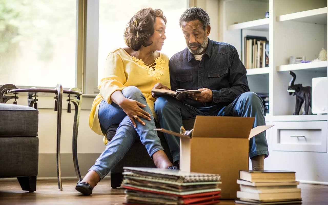 Older couple review an open book beside a moving box in their living room.
