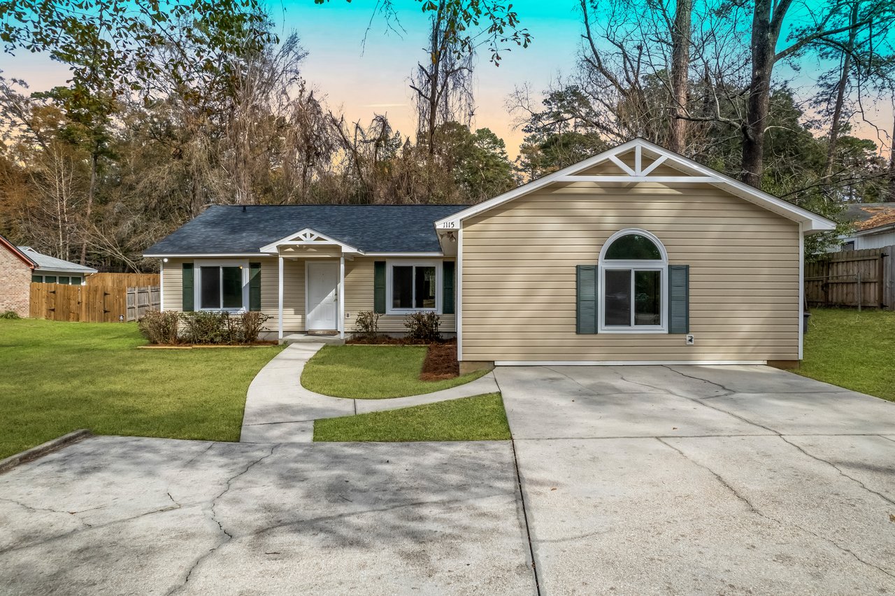 Single-story beige house with white trim and green shutters, a curved driveway leading to the entrance. Surrounded by trees under a clear sky.