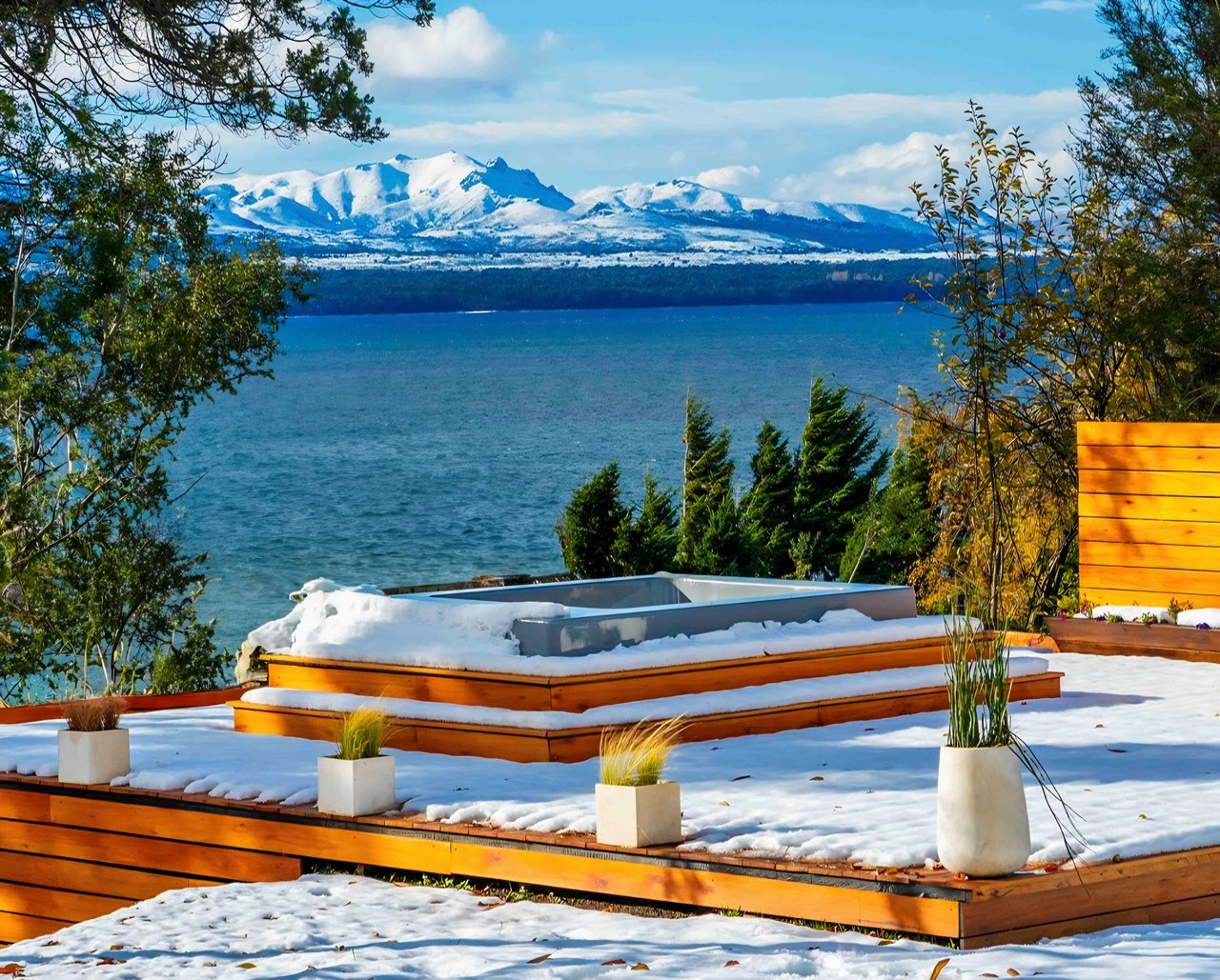 Snow-covered terrace hot tub overlooking a lake and snow-capped mountains.