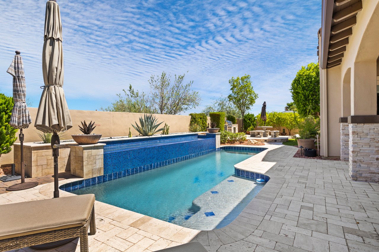 Luxury backyard patio with a swimming pool, spa, stone pavers, umbrellas, and landscaped seating area beside the house.