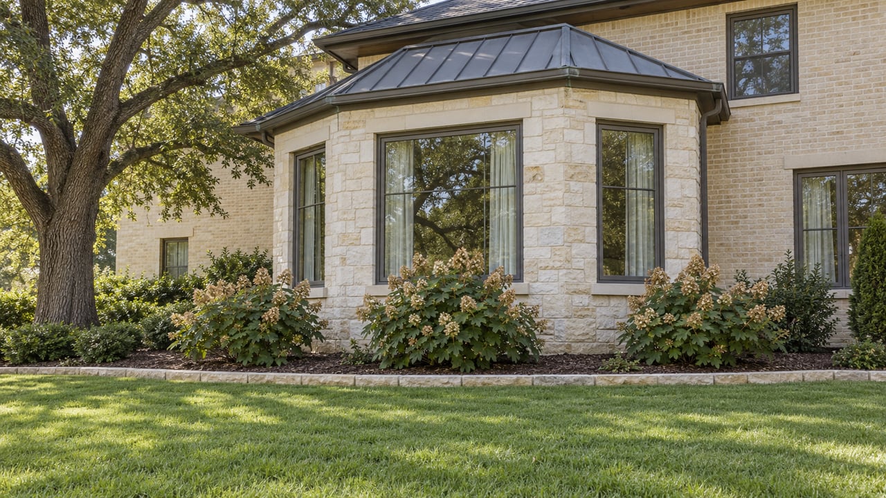 Cream limestone house with bronze-framed bay windows, a curved garden bed and a spreading live oak beside a green lawn.