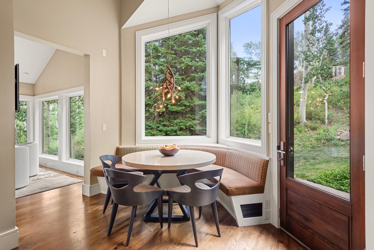 Breakfast nook with a round table, curved banquette, and large windows overlooking trees.