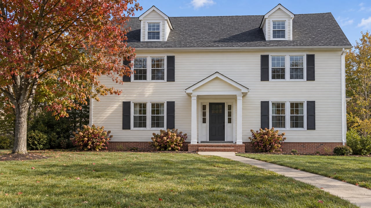 Cream-sided two-story home with paired dark shutters, a covered entry, a broad autumn lawn and a red-leaved maple.