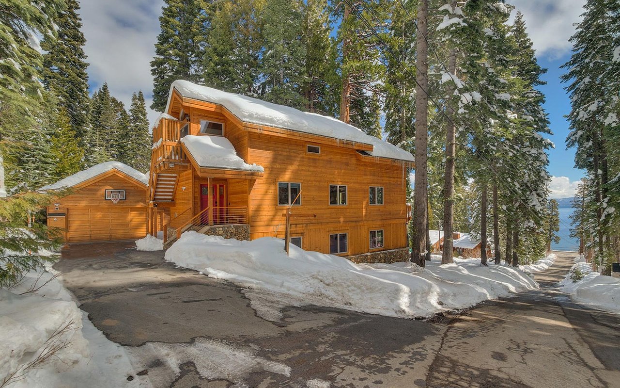 Snow-covered timber home and garage among pines, with Lake Tahoe visible beyond.