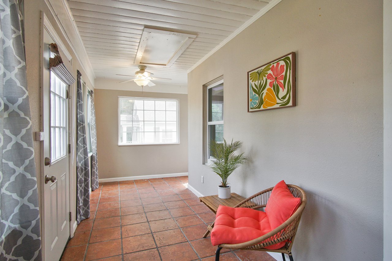 Bright sunroom with terracotta tiles, a wicker chair with a coral cushion, and a vibrant floral painting. A plant and patterned curtains add warmth.