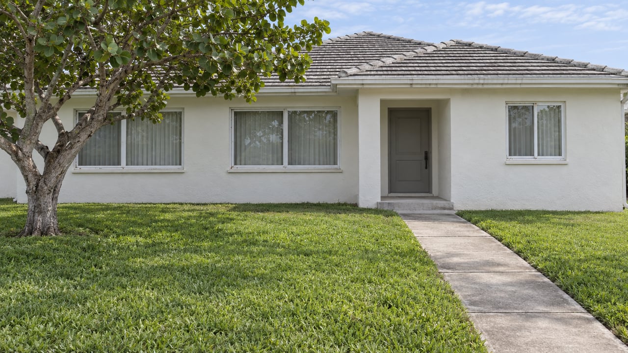 A warm-white single-story stucco home beyond a green front lawn, with a concrete entry walk and one spreading sea grape tree.
