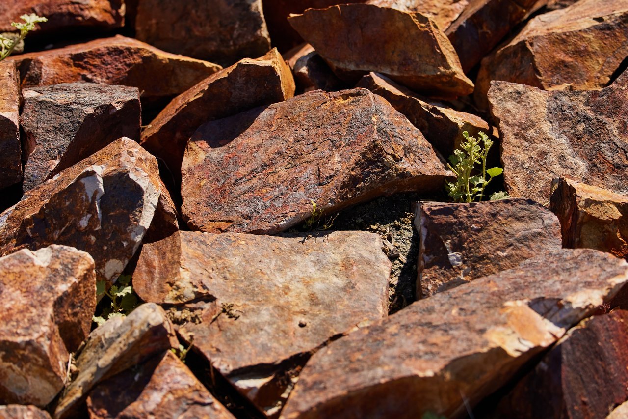 Close-up of jagged rust-colored rocks piled together in bright sunlight, with small green weeds growing between them