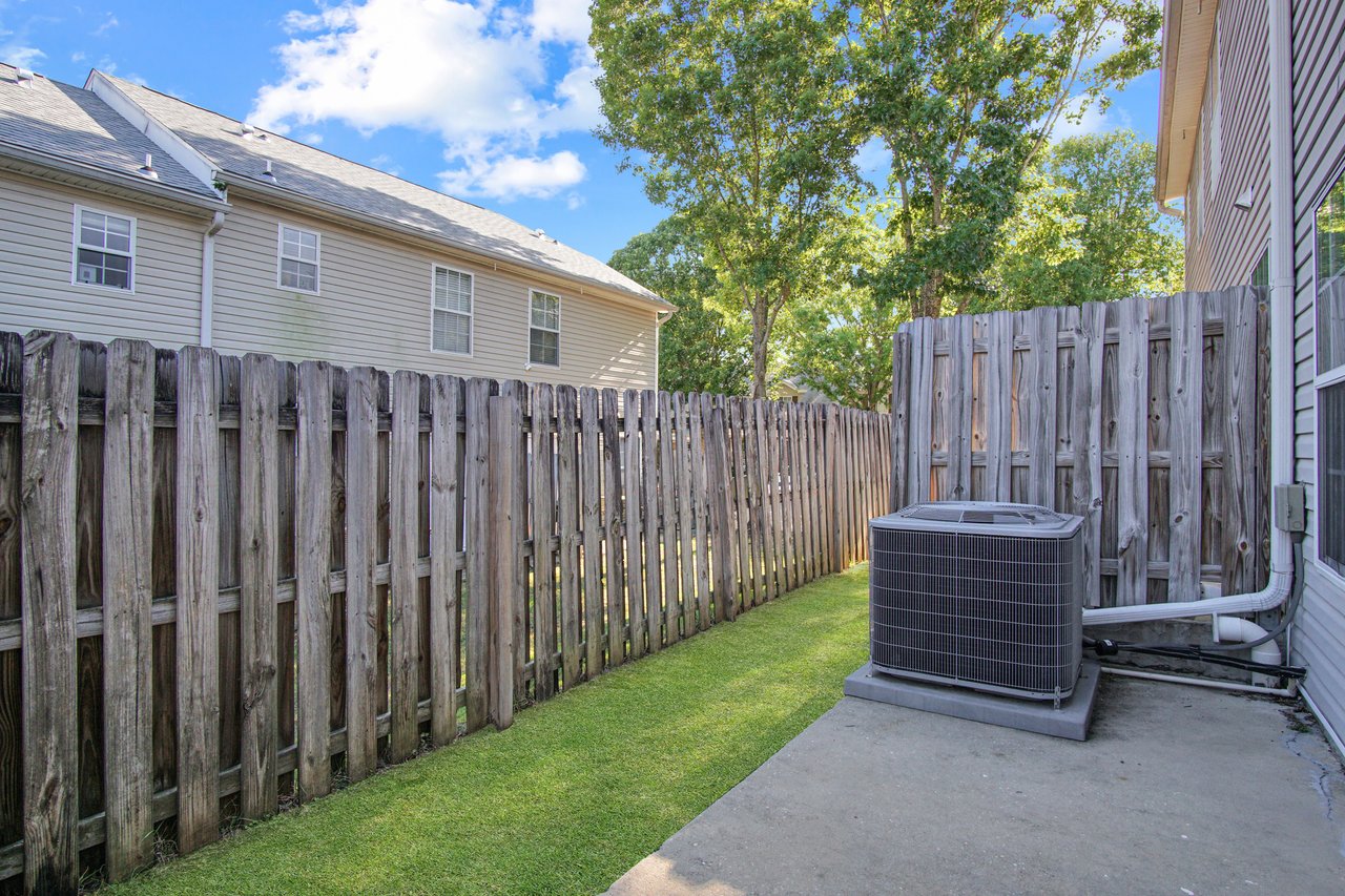 A sunlit backyard with a weathered wooden fence, green grass, and a large air conditioning unit on a concrete pad. Trees and a house are visible in the background.
