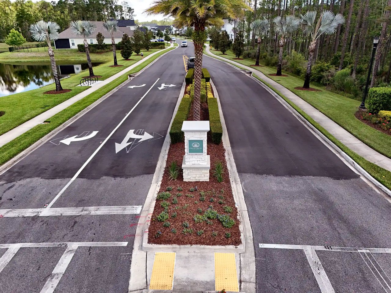 Palm Crest entrance road with a landscaped median, stone sign pillar and palms, a pond on the left and homes ahead.