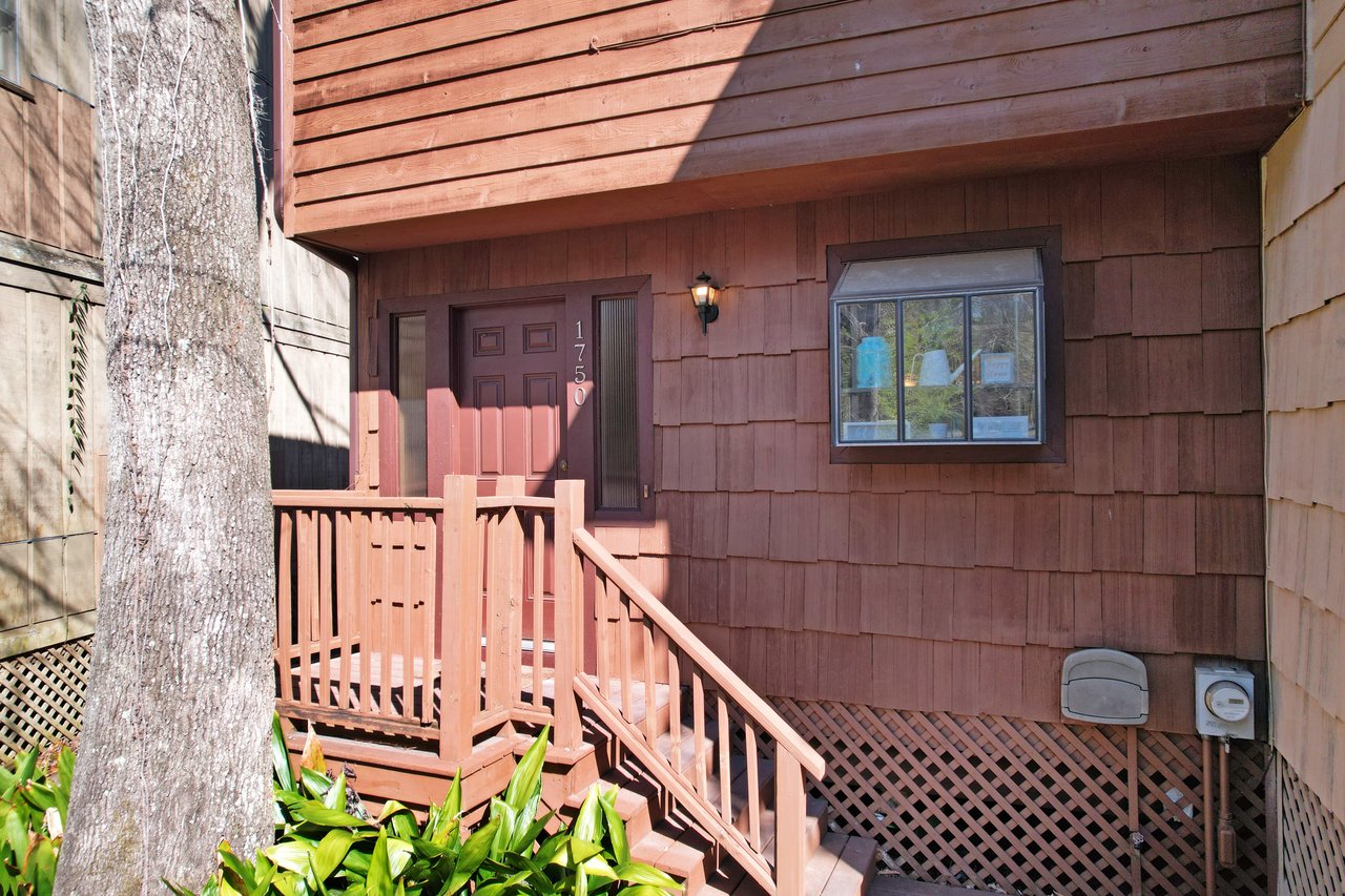 Wooden house exterior with brown siding and a small porch. The front door, marked with number 1750, is next to a window displaying plants. Cozy and rustic.