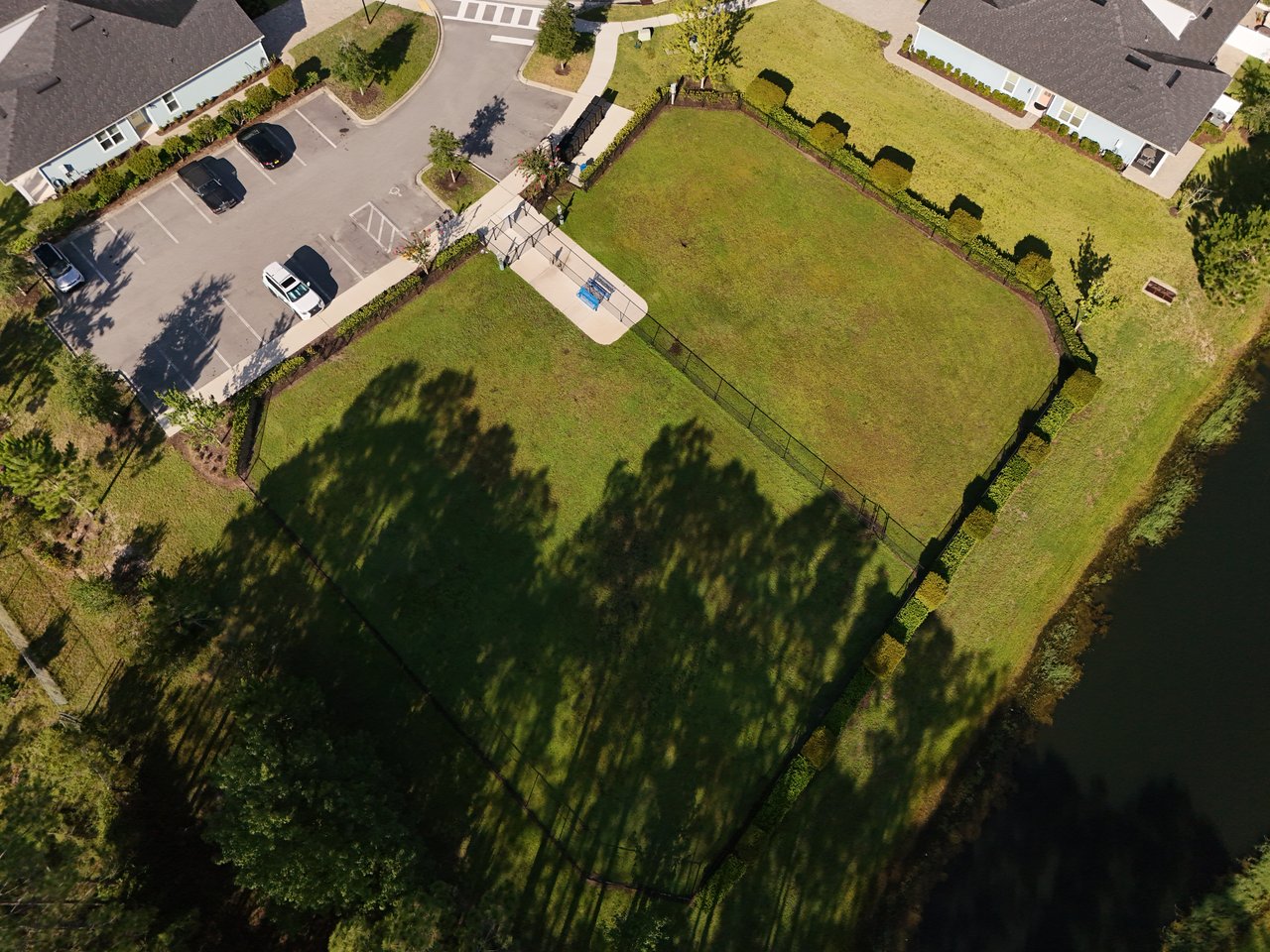 Fenced grassy recreation area beside a pond, with nearby homes and a paved seating area.