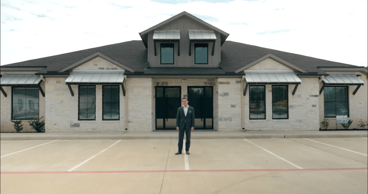 Commercial office building with a stone facade and metal awnings; a suited man stands in the parking lot.