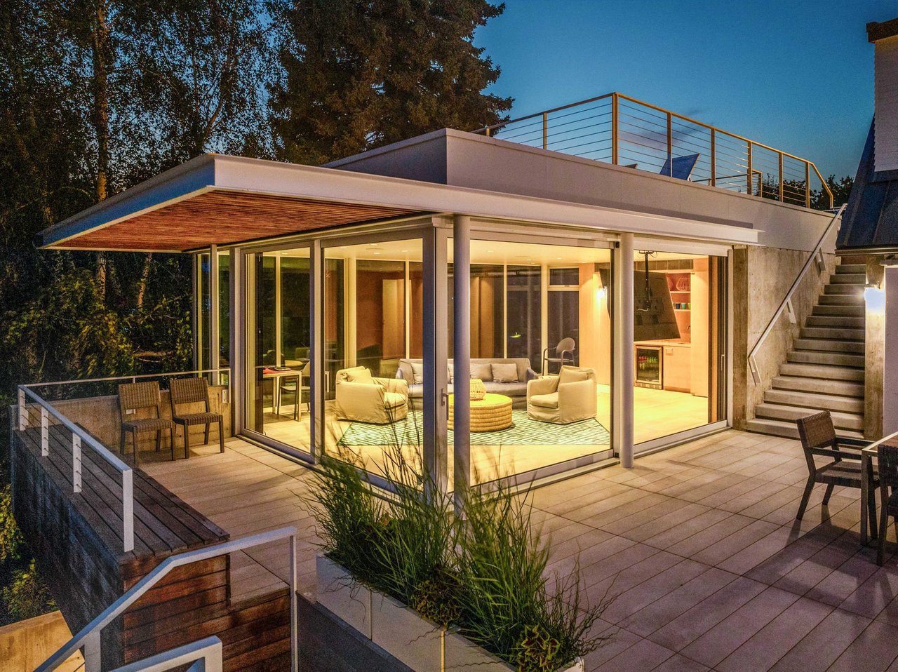A modern glass-enclosed living room with light-colored furniture, including two armchairs and a sofa, is surrounded by an outdoor patio and deck.