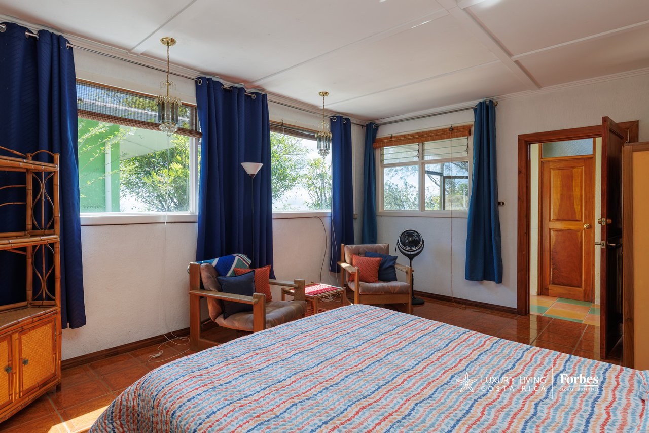 Furnished bedroom with a colorful quilted bed, navy curtains, broad windows, and wooden armchairs.