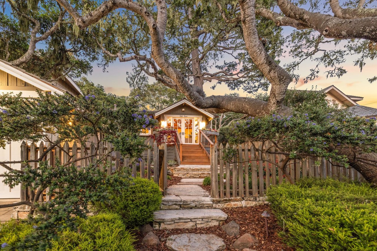 Cozy Carmel Woods cottage with lit windows under a sprawling oak tree, stone path, picket fence, and blooming garden at dusk.