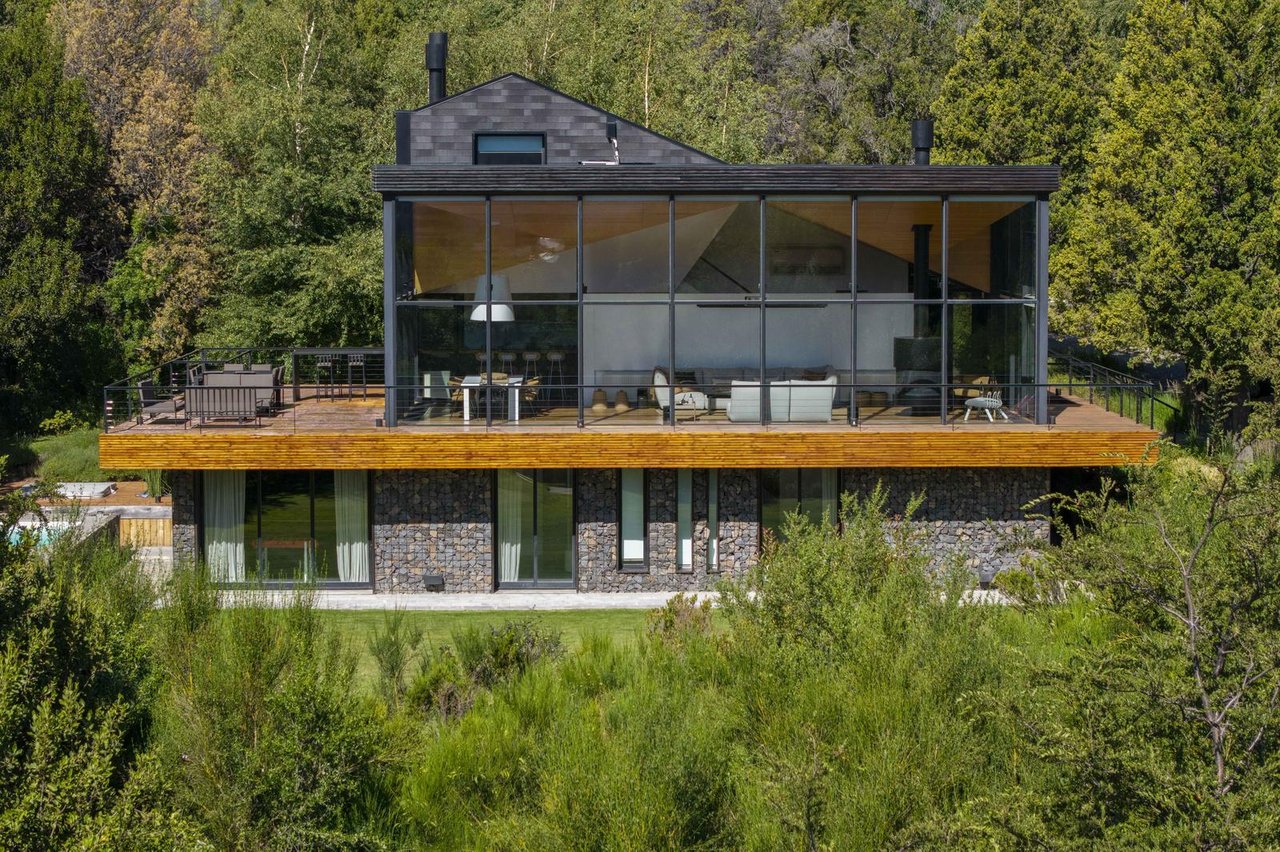 Casa Arelauquén’s glass-fronted modern home with a broad timber deck, stone walls, and wooded surroundings.