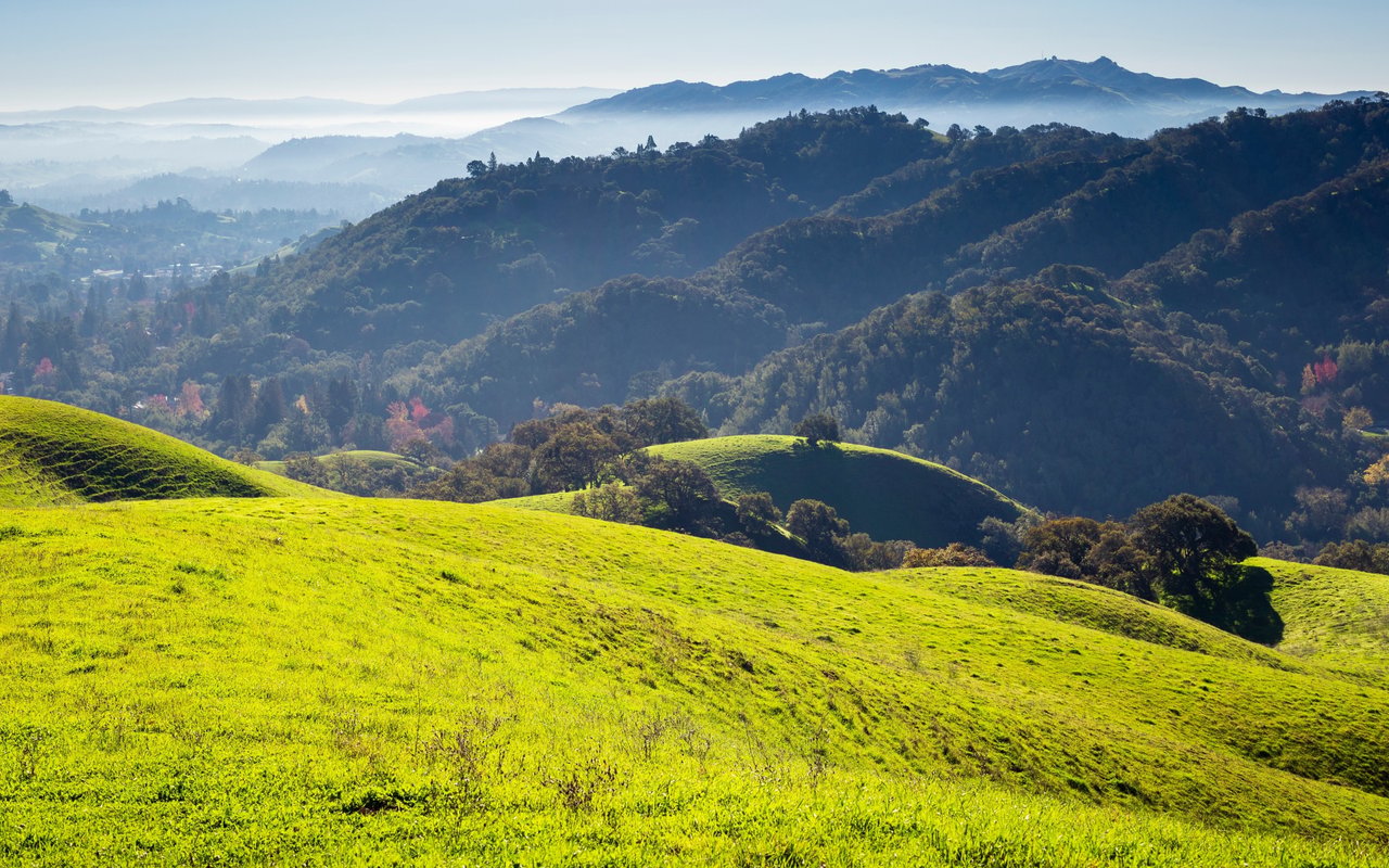 Sunlit green hills roll toward wooded ridges and hazy distant mountains.
