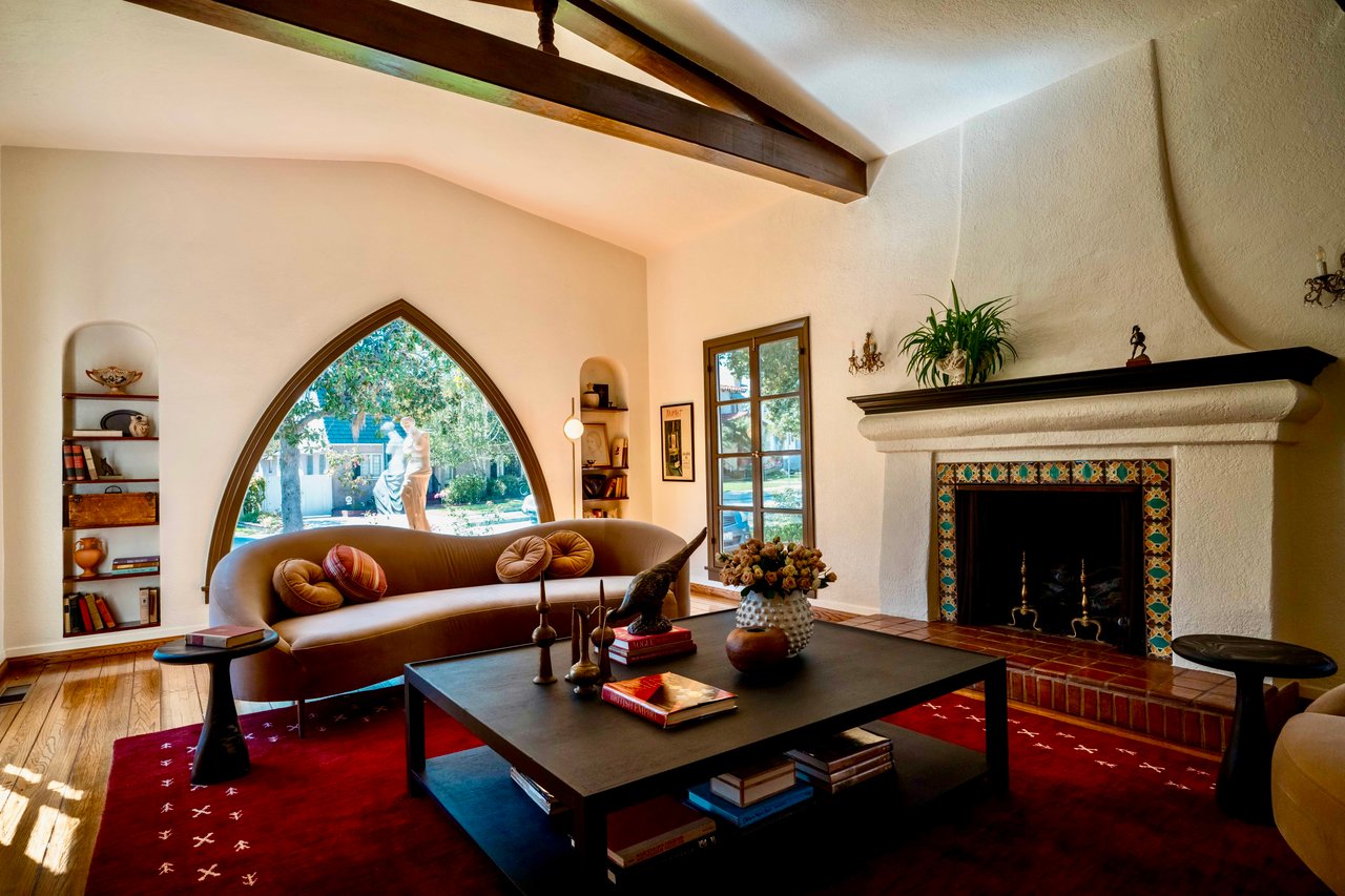 Los Angeles Spanish Revival living room with arched picture window, exposed beams, and a plaster fireplace with decorative tile; bright and staged.