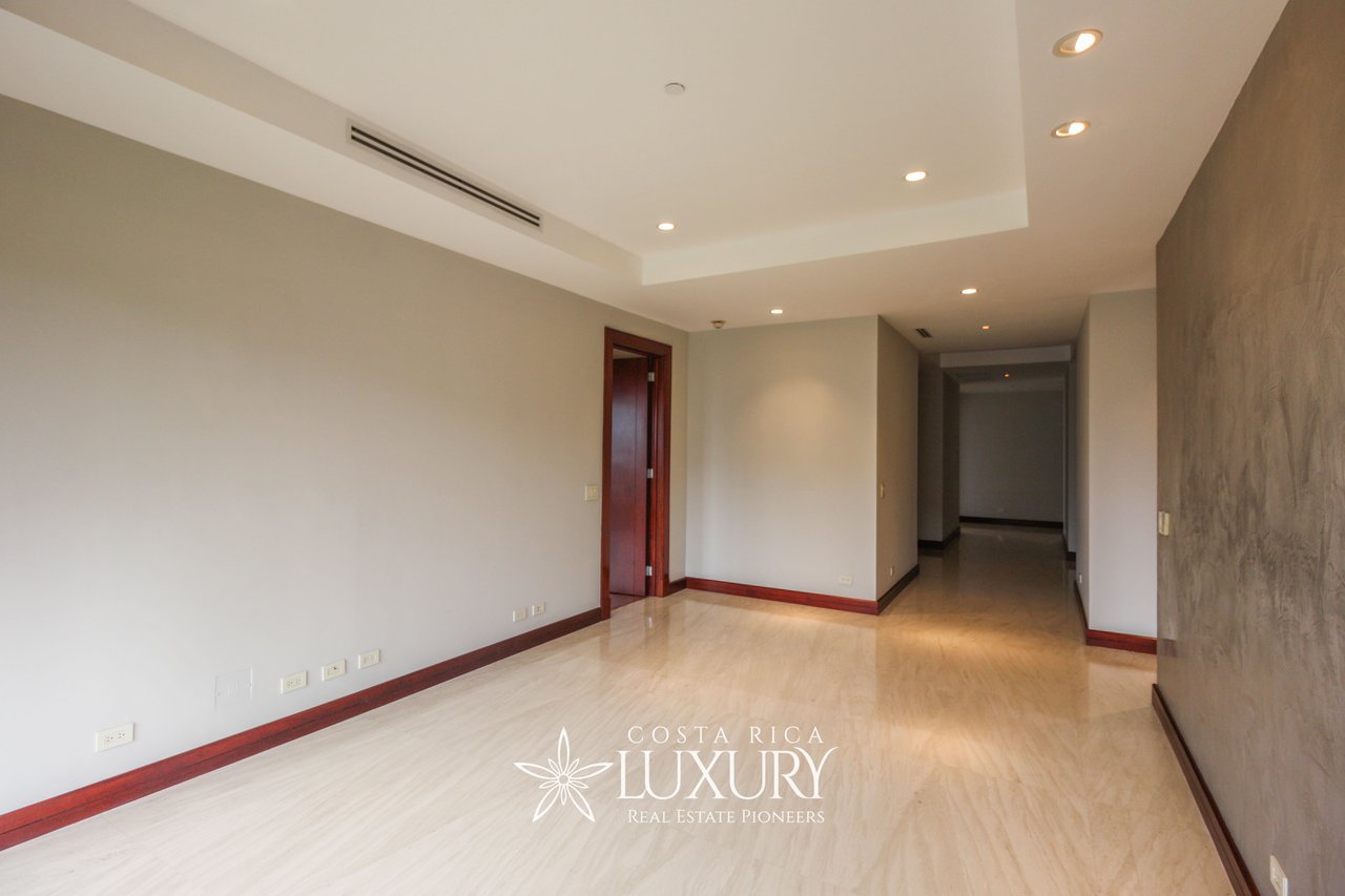 Empty apartment room with polished pale flooring, recessed lights, and a hallway.