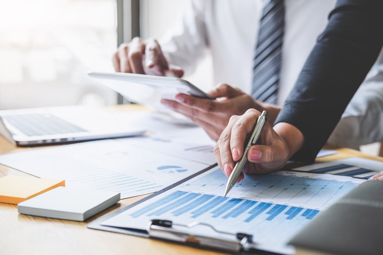 Professionals review financial charts, with one marking a blue bar graph and another holding a tablet.