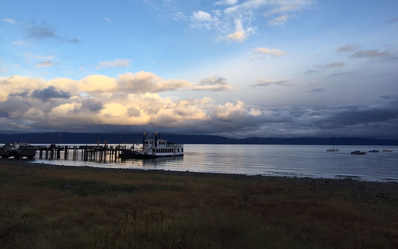 North Lake Tahoe shoreline with a pier, passenger boat, and small boats beneath dramatic clouds.