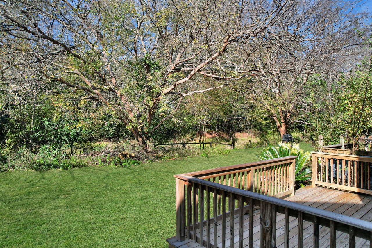 A wooden deck overlooks a lush green backyard with a large tree on the left and dense foliage. Sunlight casts shadows, creating a peaceful, natural scene.