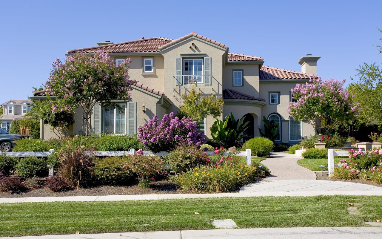 Two-story stucco home with a tile roof, balcony, and flowering landscaped front yard.