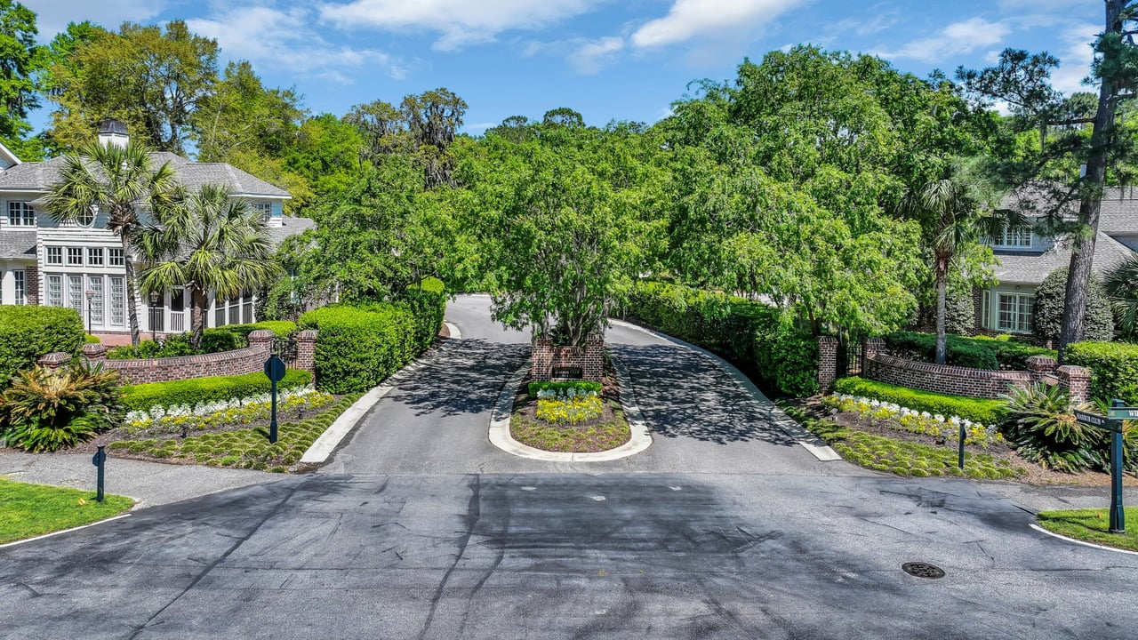 Villa neighborhood entrance road curves around a landscaped island beneath mature trees.