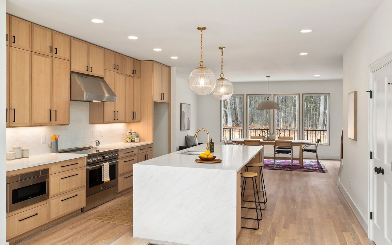 Modern kitchen with light wood cabinets, a large white island, and wooded views beyond the dining area.
